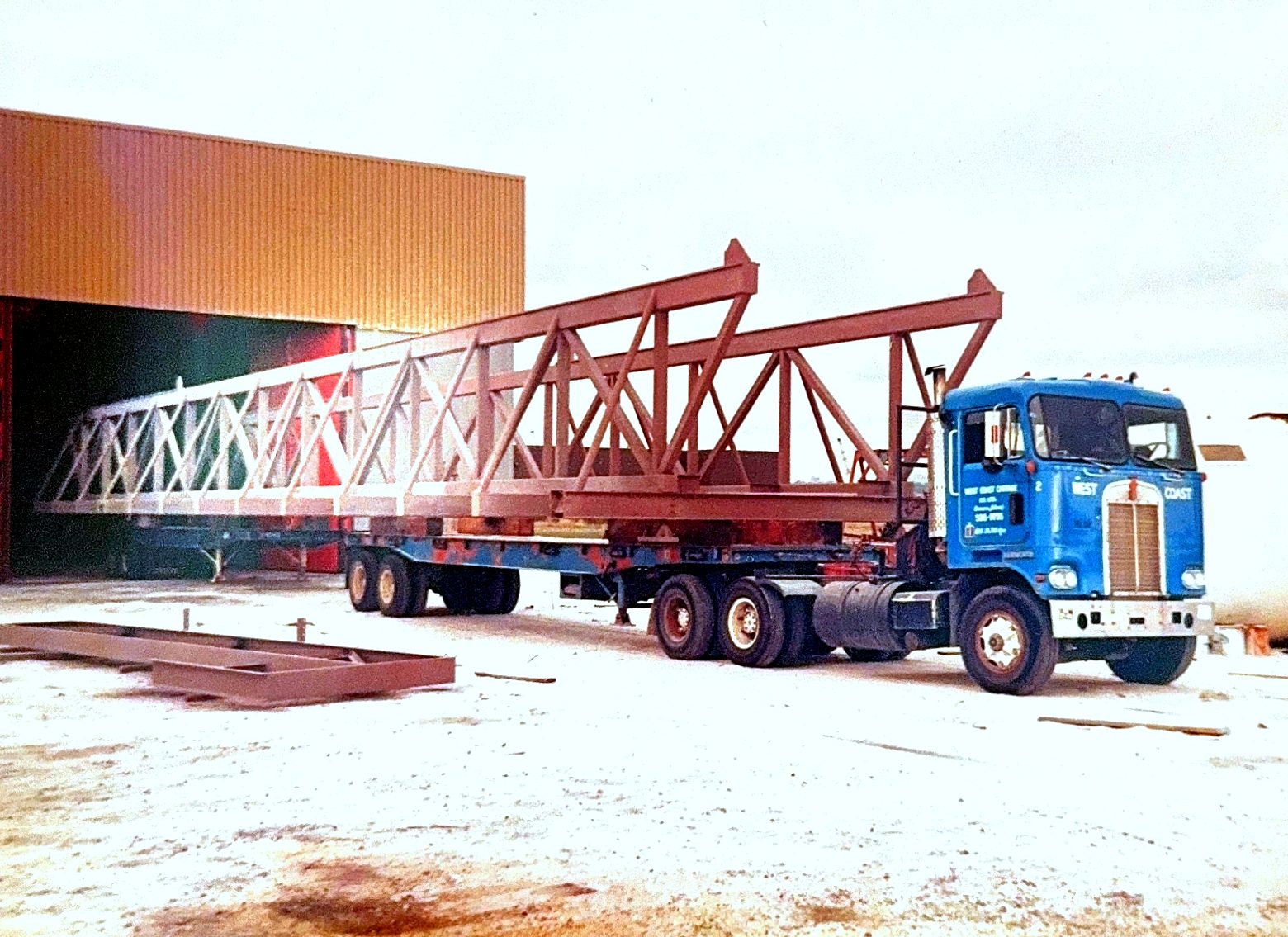 A West Coast Cartage cab-over hauling long steel roof trusses on a flat deck trailer